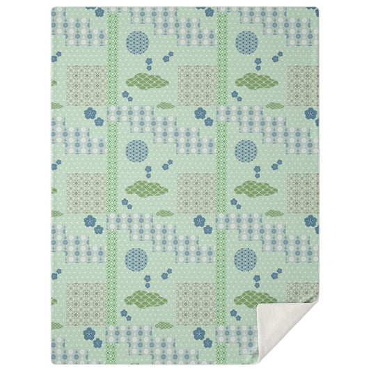 A high-angle, square photograph of a minimalist home office featuring a dark walnut wood lounge chair. Neatly draped over the chair is the Japanese Springtime Patchwork Blanket from Kaito Japan Design. The blanket features a sophisticated patchwork of soft Sage Green, muted Sakura Pink, and Cream tones, with traditional Wagara motifs like stylized blossoms and geometric patterns. Natural morning light streams through a window, creating a calm, professional atmosphere.