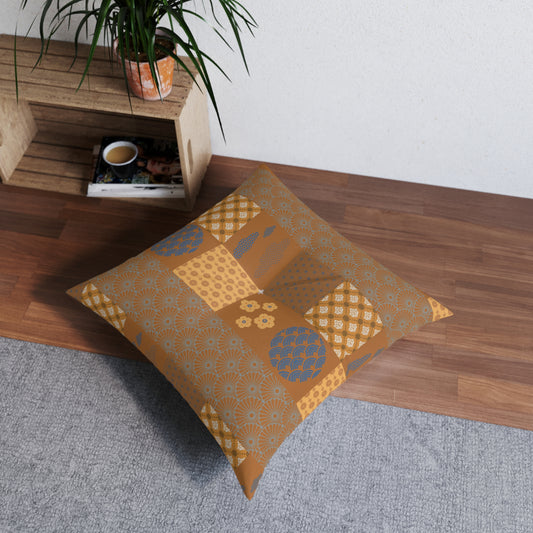 A square floor pillow with a patchwork pattern in mustard yellow, tan, and blue, resting on a gray rug next to a wooden shelf with a potted plant.