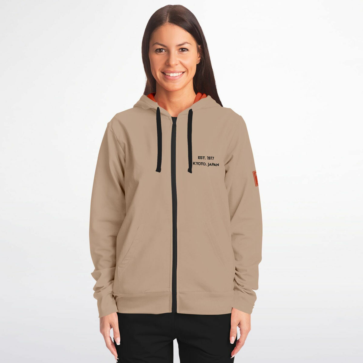 A woman modeling a beige zip-up hoodie with black drawstrings, a black front zipper, and black text on the left chest reading 'EST. 1997 KANSAS CITY, MO'.