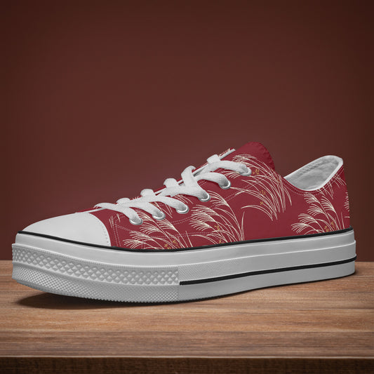 A low-top canvas sneaker with a red upper featuring a white palm leaf pattern, white laces, a white rubber toe cap, and a thick white platform sole with a black stripe, resting on a wooden surface against a solid dark brown background.