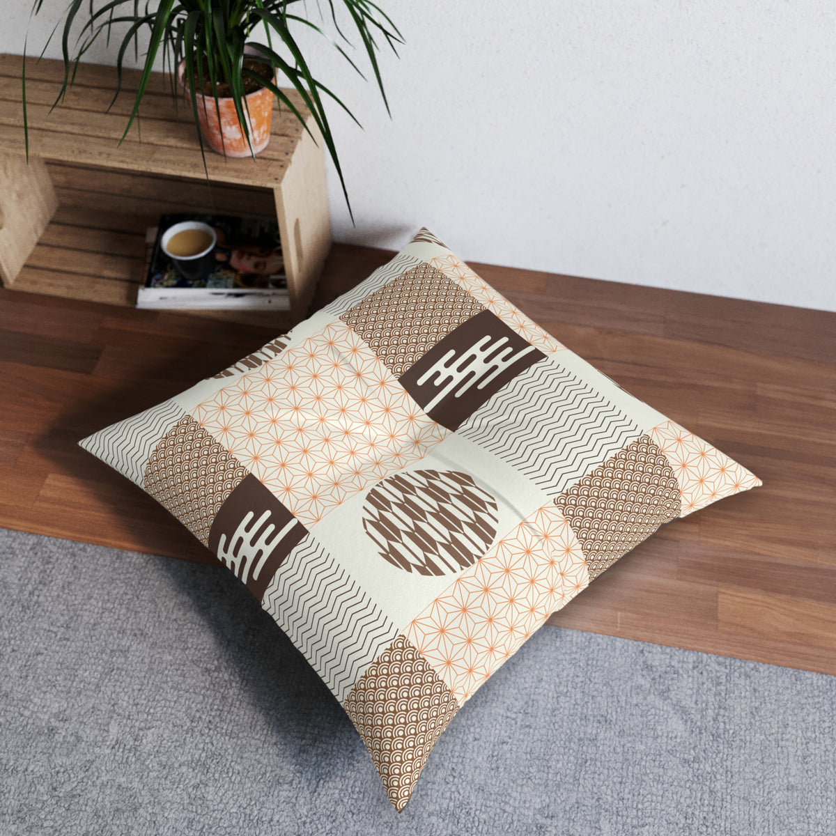 A square decorative throw pillow with a patchwork of beige, brown, and orange geometric patterns, resting on a gray rug next to a wooden shelf with a potted plant.