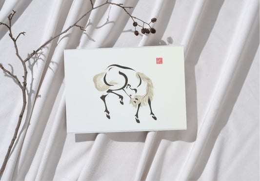 A white art print featuring a Japanese ink painting by Aoyama Seizan, depicting a horse in a dynamic, scratching pose with fluid black and light gray brushstrokes. A red square seal with Japanese characters is in the upper right corner of the print. The print lies on a draped white fabric surface, with a bare tree branch featuring small seed pods extending across the top left. Natural light casts shadows, adding depth to the serene composition