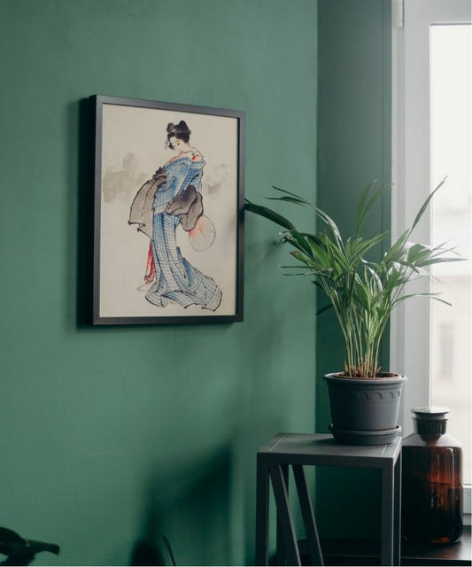 Framed artwork of a woman in a blue dress on a green wall with a plant and bottle in the foreground.