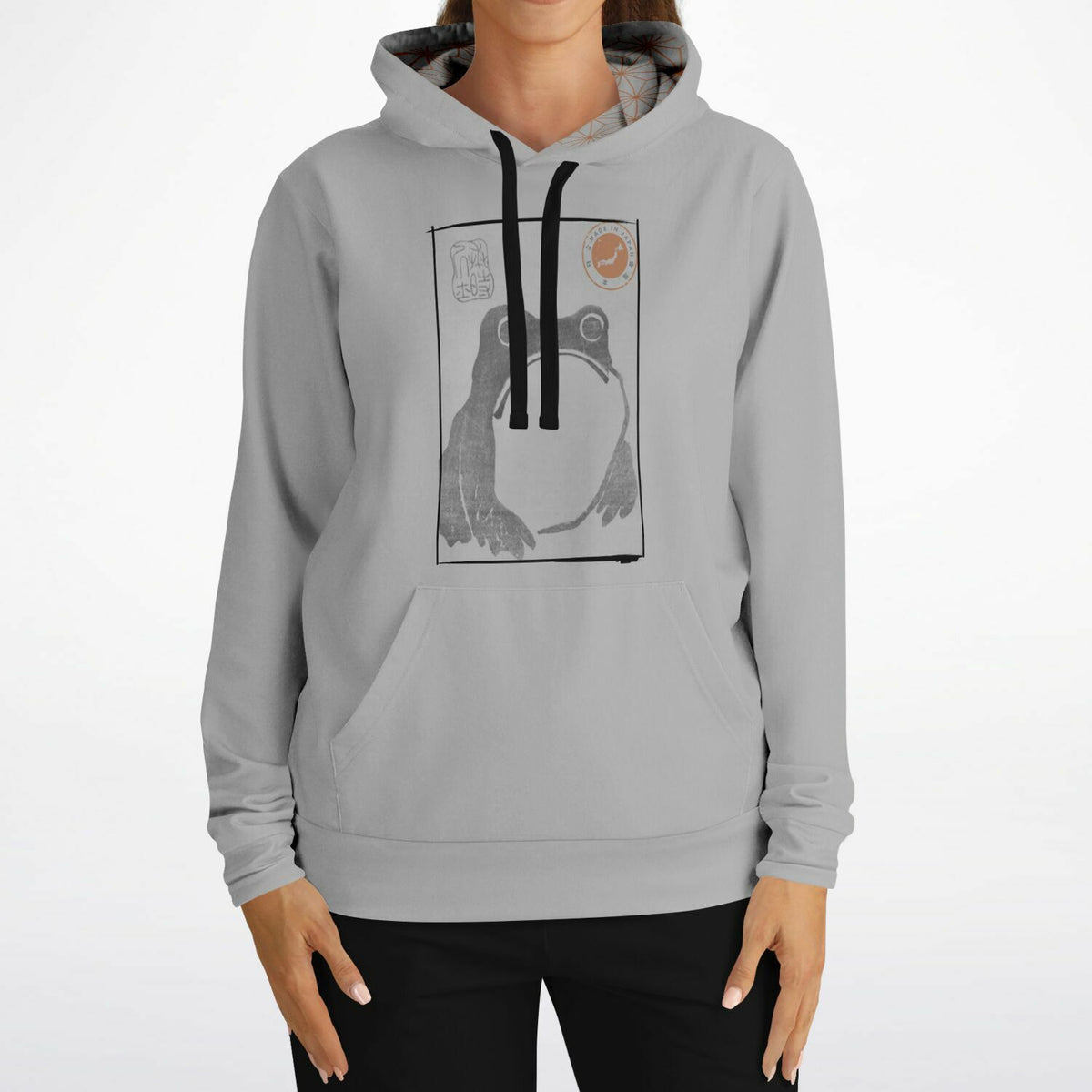 A woman wearing a light gray pullover hoodie with a black and white graphic print of a frog on the chest, paired with black pants, against a plain white background.