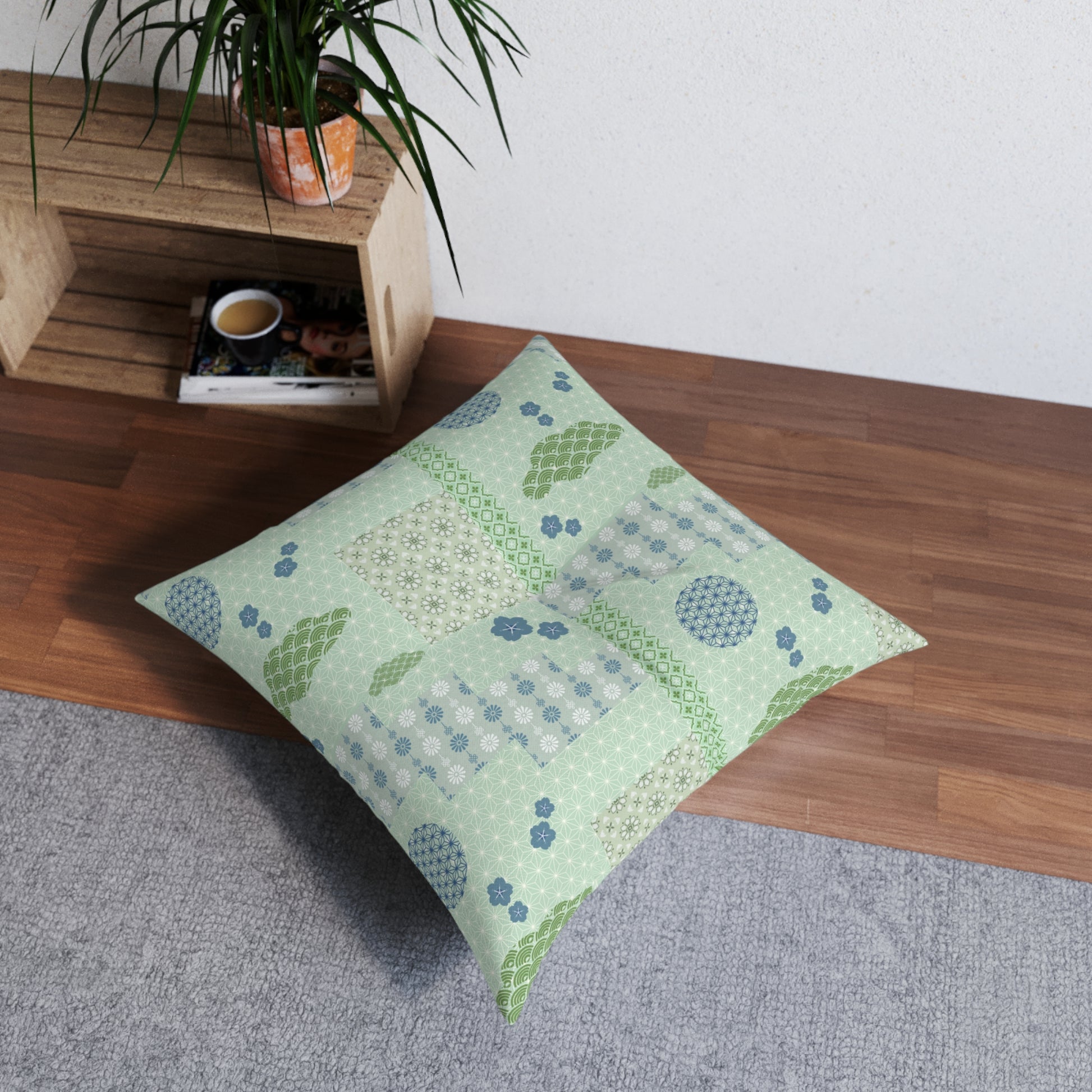 A square floor pillow with a patchwork pattern in sage green, light blue, and white, styled on a gray rug next to a wooden shelf with a potted plant.