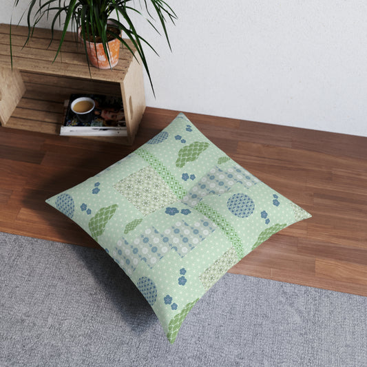 A square floor pillow with a patchwork pattern in sage green, light blue, and white, styled on a gray rug next to a wooden shelf with a potted plant.