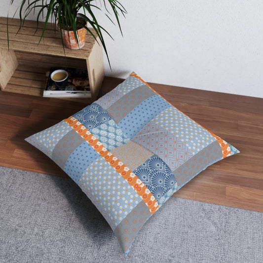 A square patchwork floor pillow with a Japanese-inspired patterned design in shades of blue, gray, and orange, styled on a wooden floor next to a potted plant.