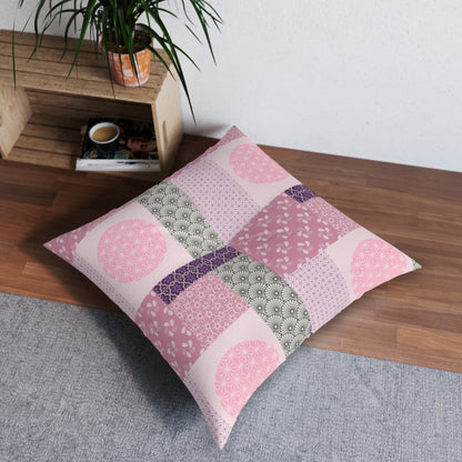 A square patchwork throw pillow in shades of pink, purple, and sage green, styled on a gray rug next to a wooden shelf with a potted plant.