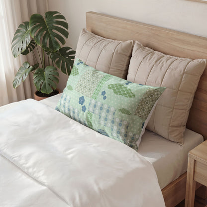 A decorative rectangular lumbar pillow with a green and blue patchwork floral pattern rests on a bed with white bedding, flanked by two beige quilted pillows and a wooden headboard, with a potted monstera plant in the background.