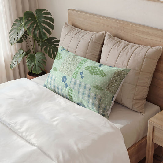 A decorative rectangular lumbar pillow with a green and blue patchwork floral pattern rests on a bed with white bedding, flanked by two beige quilted pillows and a wooden headboard, with a potted monstera plant in the background.