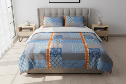 A bed dressed in a patchwork-style blue and gray patterned comforter with two matching pillow shams, styled in a bright bedroom with light wood nightstands and a gray upholstered headboard.
