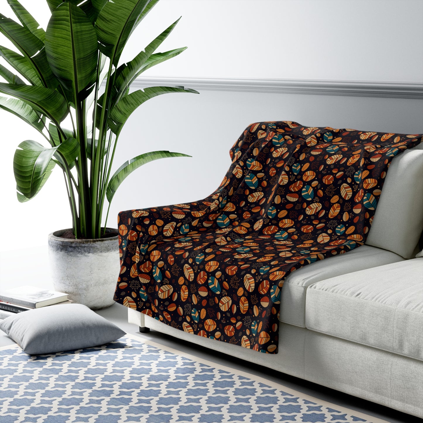 A dark throw blanket with an orange, brown, and teal leaf pattern draped over a light gray sofa in a modern living room setting with a potted plant and patterned rug.
