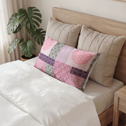 A rectangular patchwork pillow in shades of pink, purple, and green is styled on a bed with white linens, flanked by two beige pillows and a wooden headboard, with a green monstera plant in the background.