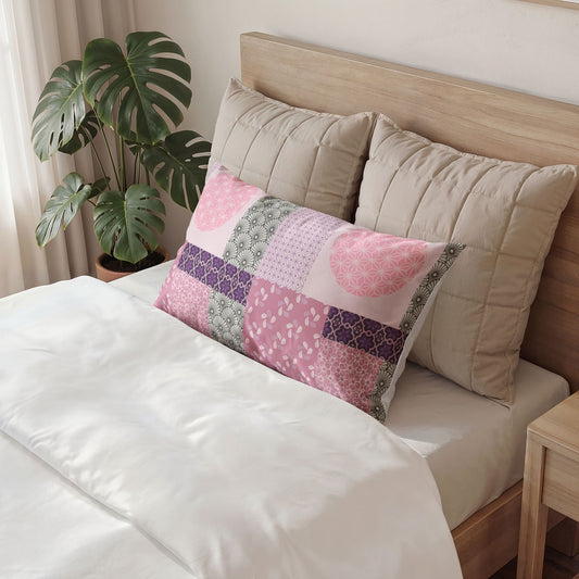 A rectangular patchwork pillow in shades of pink, purple, and green is styled on a bed with white linens, flanked by two beige pillows and a wooden headboard, with a green monstera plant in the background.