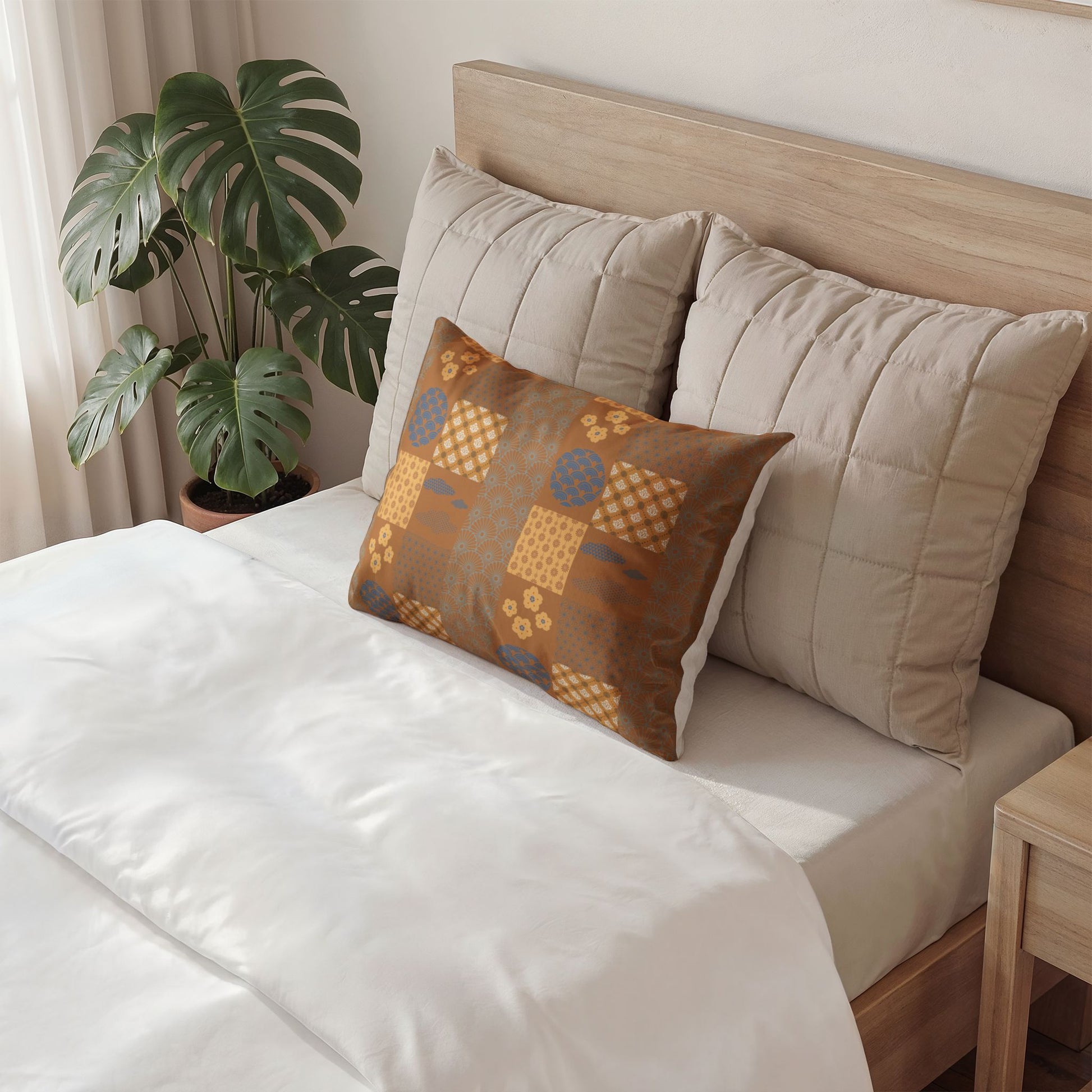 A styled bedroom scene showing a bed with white bedding, two large beige quilted pillows, and a smaller square decorative throw pillow in brown, mustard, and blue geometric patterns. A potted monstera plant sits beside the bed.