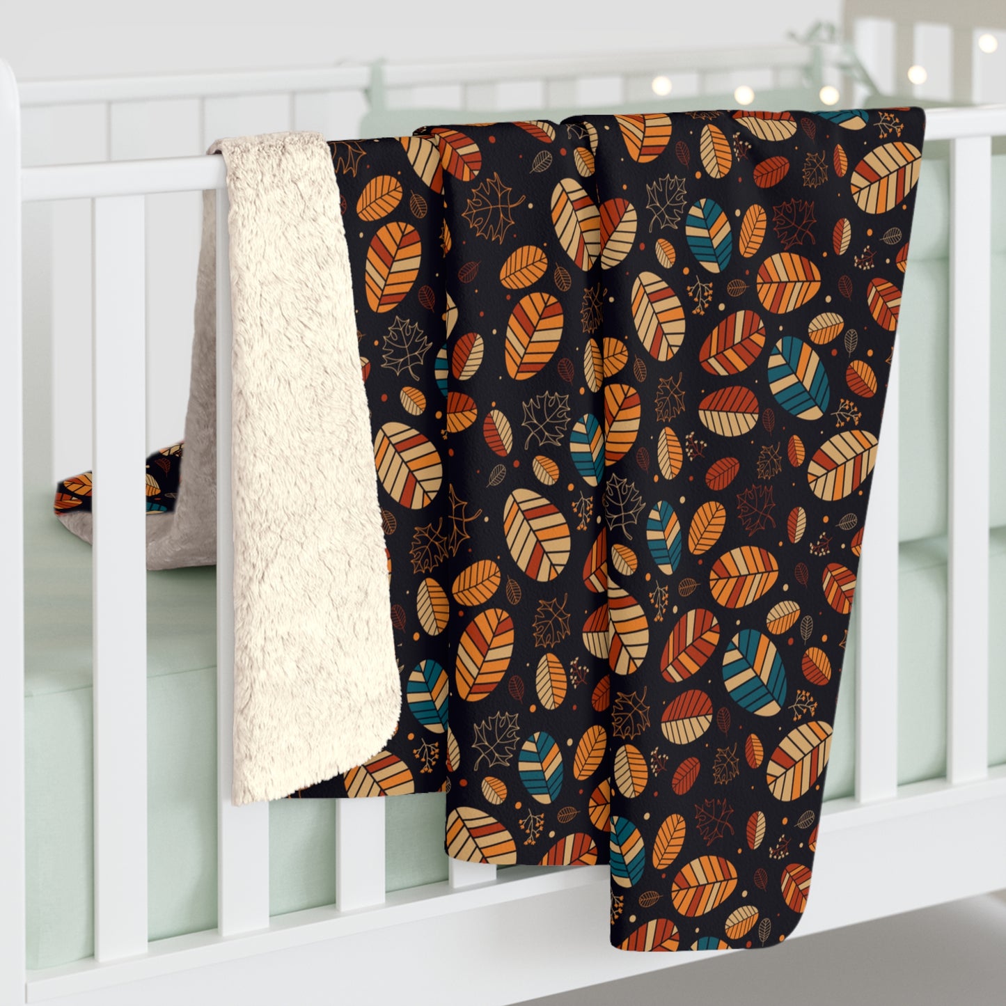 A baby blanket draped over a white crib, featuring a dark background with an autumn leaf pattern in orange, beige, and teal, and a soft cream sherpa lining.