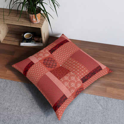 A square decorative throw pillow with a patchwork of red, orange, and beige Japanese-style patterns, styled on a gray rug next to a wooden bench with a potted plant.