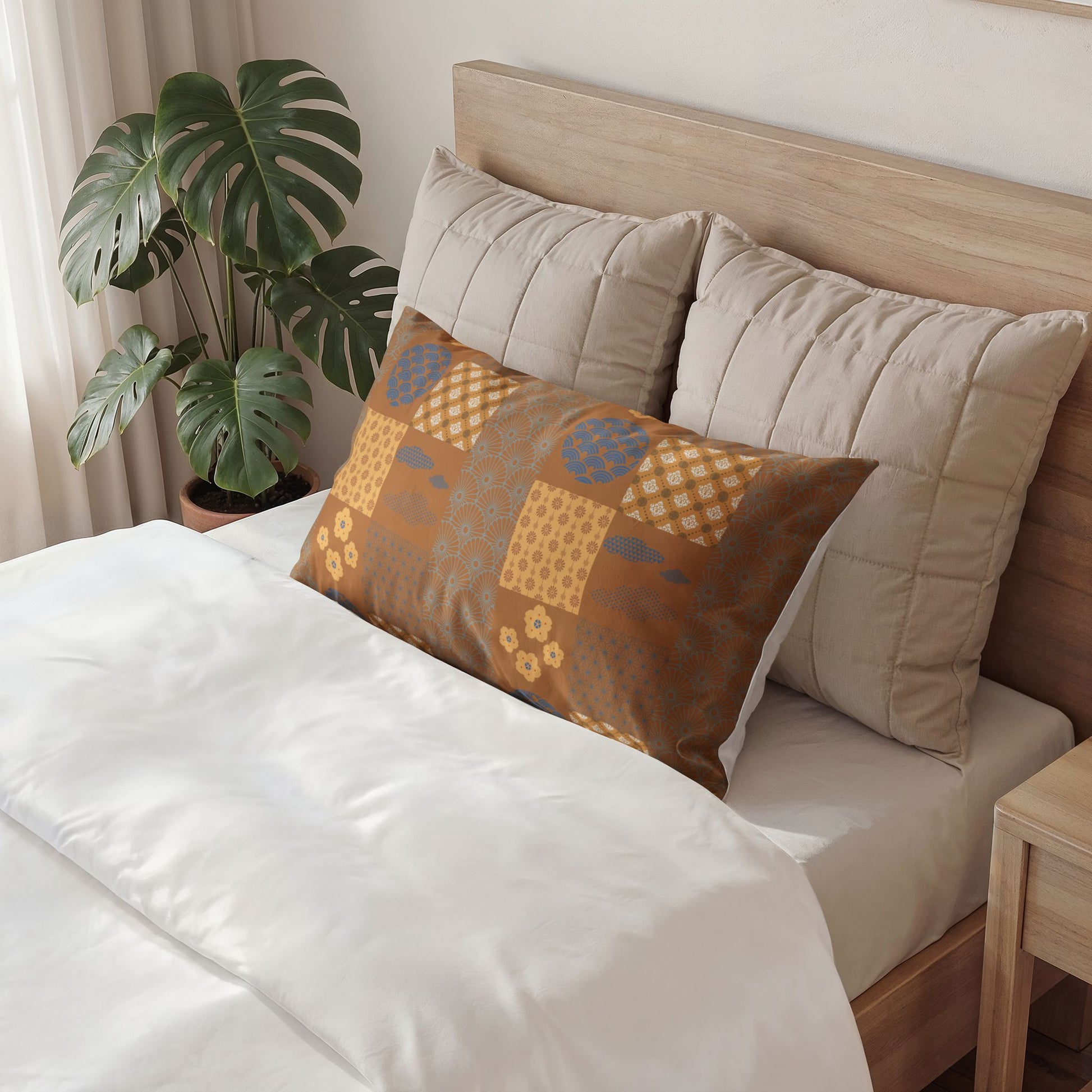 A decorative rectangular throw pillow with a patchwork pattern in brown, beige, and blue, styled on a bed with white linens and beige pillows in a bedroom setting.