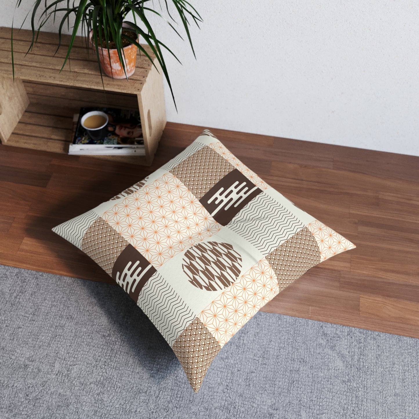 A square decorative throw pillow with a patchwork of Japanese-inspired geometric patterns in beige, brown, and white, styled on a gray rug next to a wooden shelf with a potted plant.