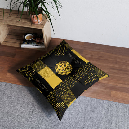 A square floor pillow with a black and yellow geometric patchwork pattern, styled on a gray rug next to a wooden bench with a potted plant.