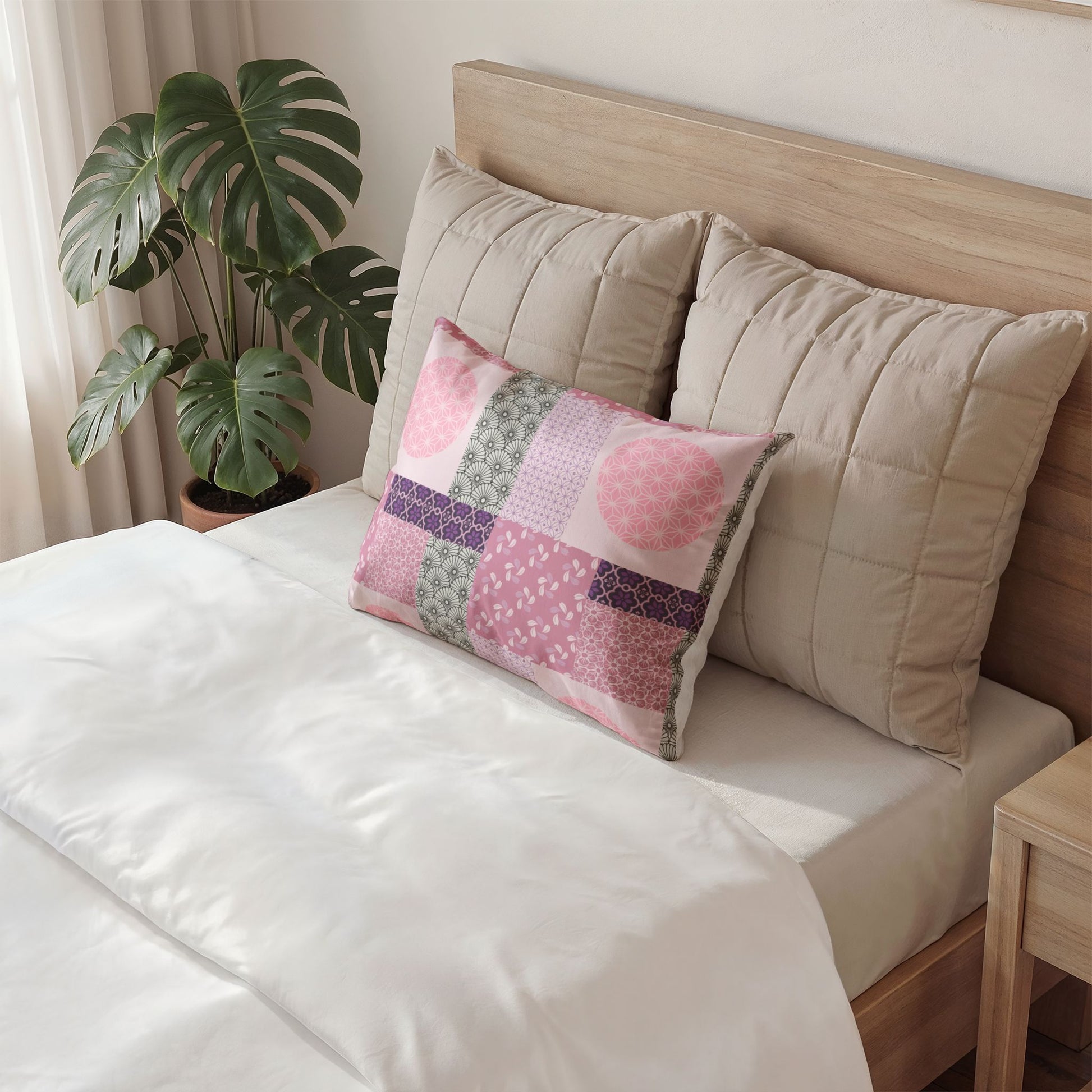 A decorative rectangular throw pillow with a red and cream patchwork pattern sits on a bed with white bedding, flanked by two large beige quilted pillows against a light wood headboard. A potted monstera plant is visible in the background.