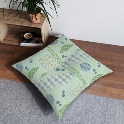 A square decorative throw pillow with a patchwork print in sage green, light blue, and white, styled on a wooden bench in a home setting next to a potted plant.