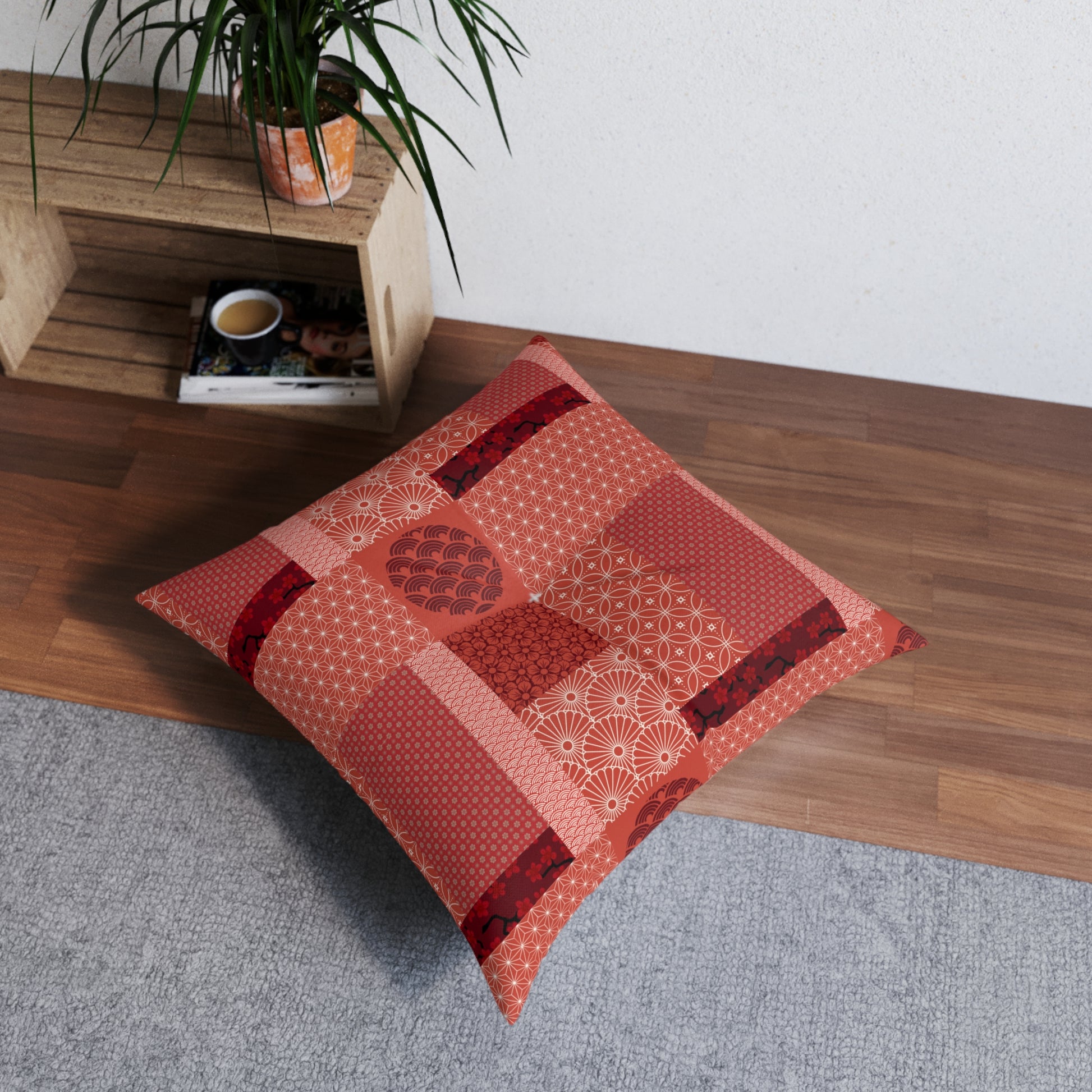 A square patchwork floor pillow in shades of red, orange, and pink with various Japanese-inspired patterns, styled on a gray rug next to a wooden shelf with a plant.