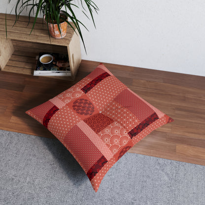 A square patchwork floor pillow in shades of red, orange, and pink with various Japanese-inspired patterns, styled on a gray rug next to a wooden shelf with a plant.