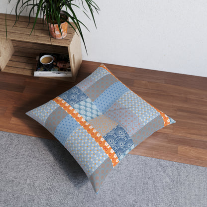 A square decorative floor pillow with a patchwork fabric design in shades of blue, gray, and orange, styled on a gray rug next to a wooden shelf with a potted plant.