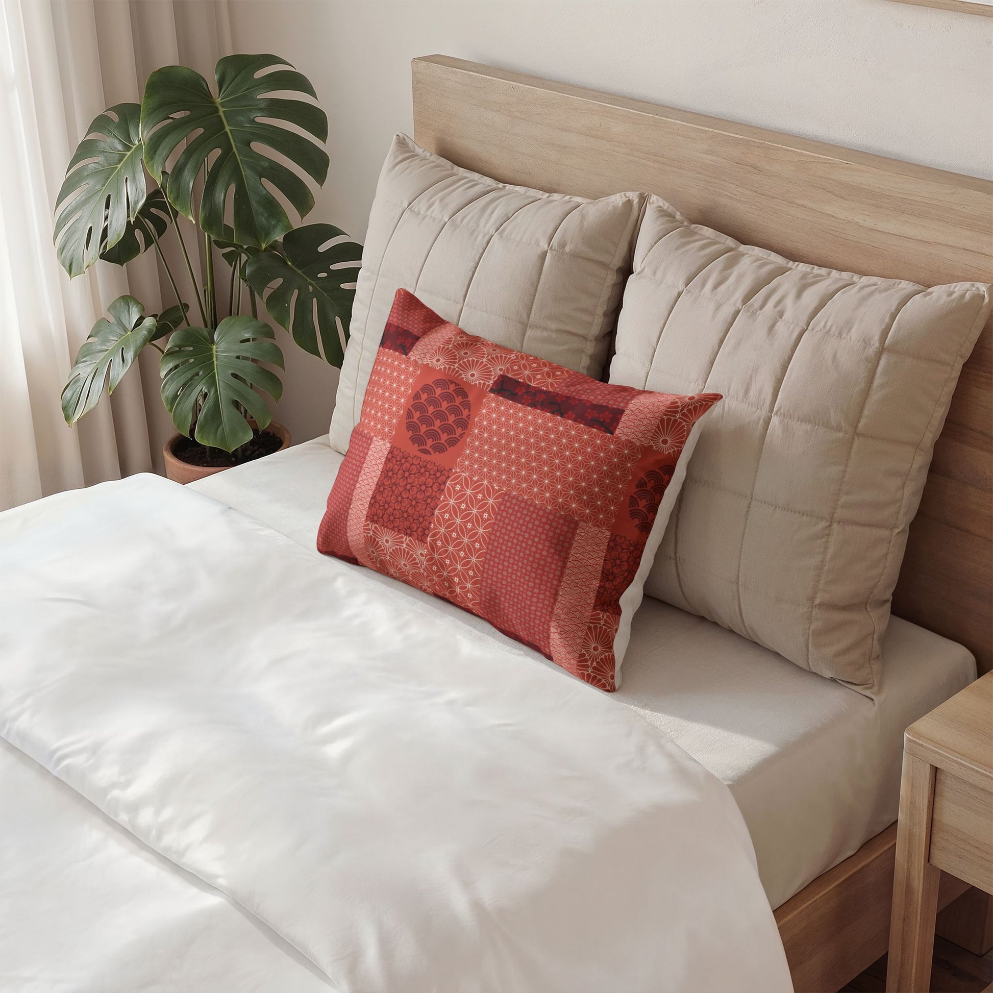 A styled bedroom scene showing a bed with white bedding, two large beige quilted pillows, and a smaller square decorative throw pillow in brown, mustard, and blue geometric patterns. A potted monstera plant sits beside the bed.