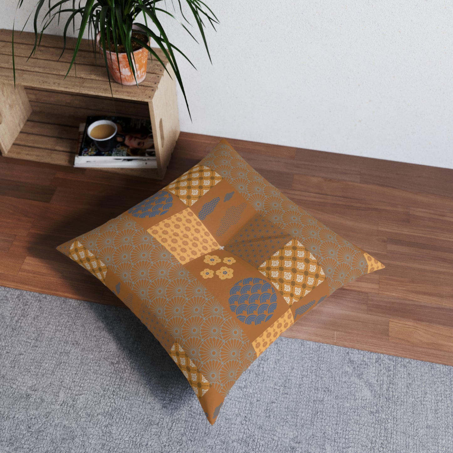 A square floor pillow with a patchwork pattern in mustard yellow, tan, and blue, resting on a gray rug next to a wooden shelf with a potted plant.