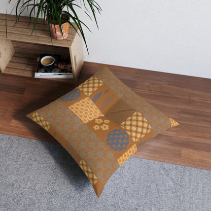 A square floor pillow with a patchwork pattern in mustard yellow, tan, and blue, resting on a gray rug next to a wooden shelf with a potted plant.