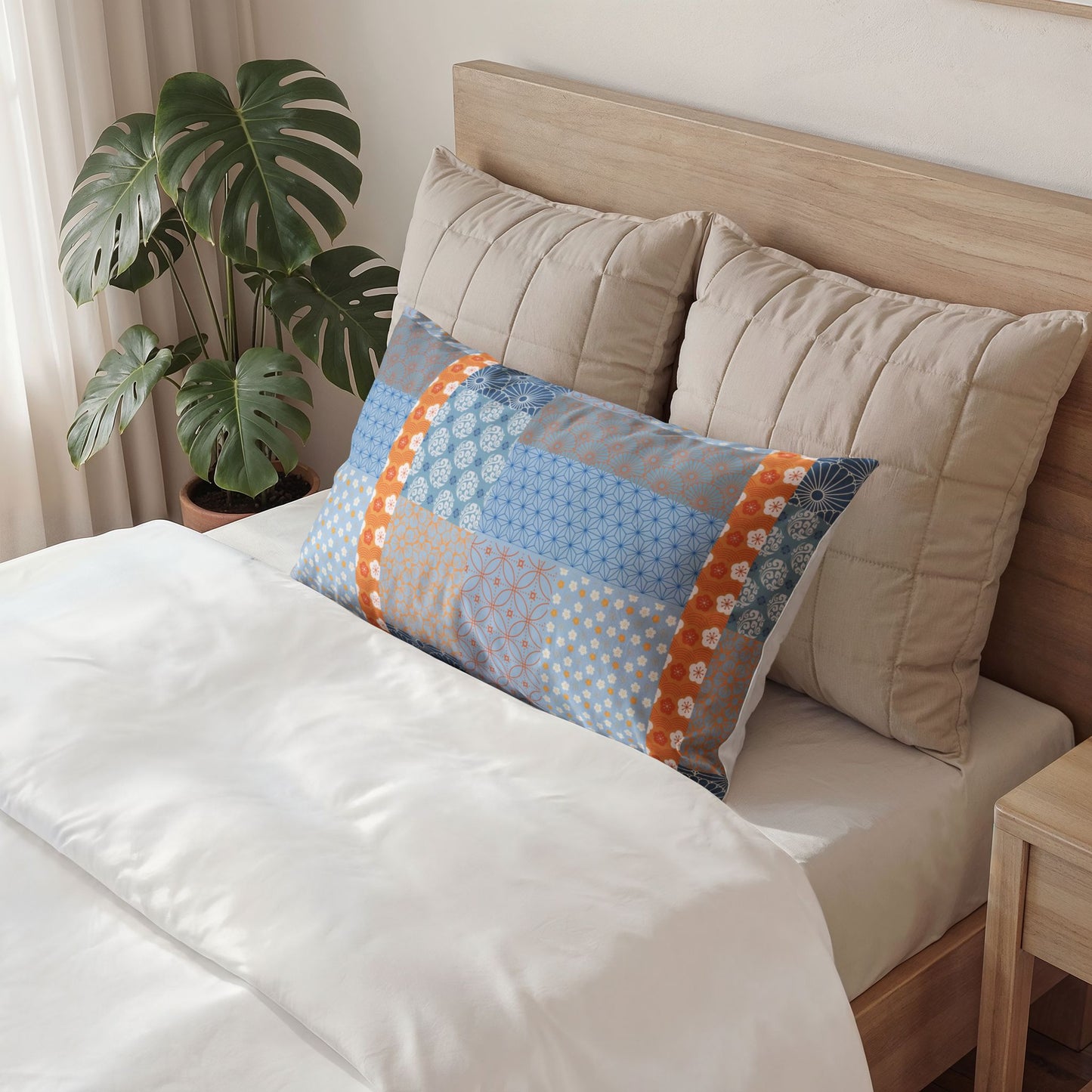 A decorative rectangular lumbar pillow with a blue, orange, and white patchwork pattern sits on a bed with white bedding and beige pillows, next to a potted green plant.