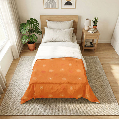 An orange quilted comforter with a subtle sunburst pattern is styled on a bed in a bright, modern bedroom. The bed is neatly made with white sheets and pillows, set against a light wood headboard, with a matching wooden nightstand, potted green plants, and a woven rug on the floor.