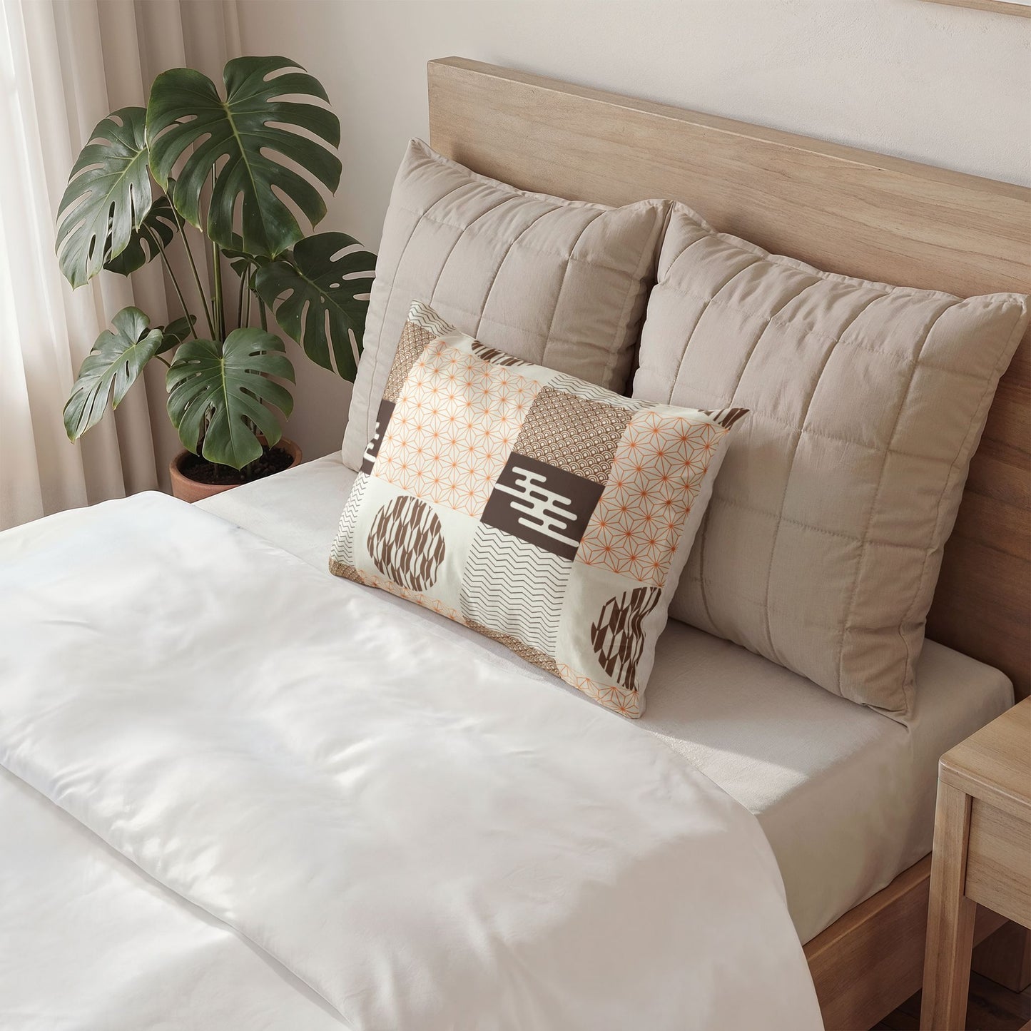 A styled bedroom scene showing a bed with white bedding, two large beige quilted pillows, and a smaller rectangular patchwork accent pillow in cream, beige, brown, and orange tones. A wooden headboard, side table, and potted monstera plant are visible.