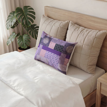 A decorative rectangular throw pillow with a purple and lavender patchwork quilted pattern, styled on a bed with beige pillows and a wooden headboard next to a potted green plant.
