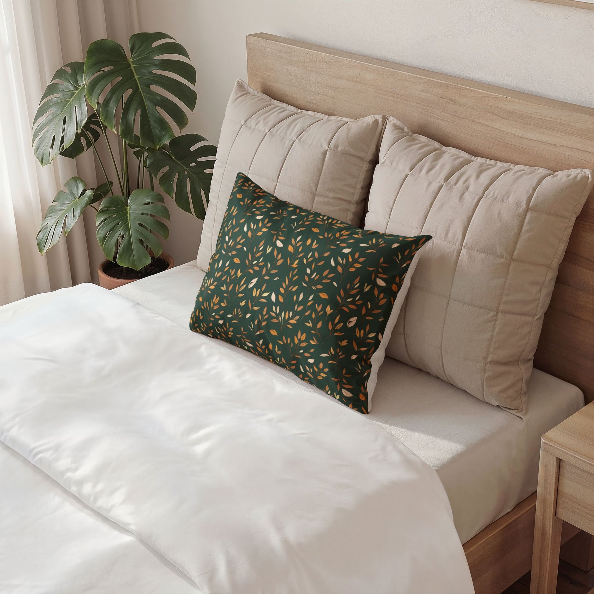A styled bedroom scene showing a bed with white bedding, two large beige quilted pillows, and a smaller dark green decorative throw pillow with a subtle circular pattern. A wooden headboard, plant, and bedside table are visible.