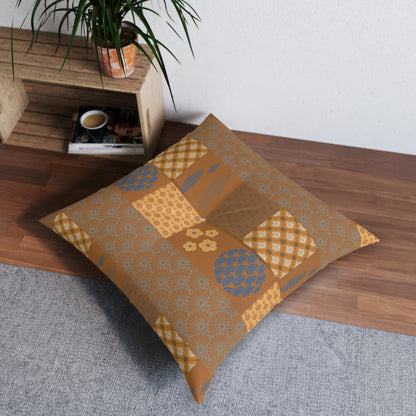 A square decorative throw pillow with a patchwork-style geometric pattern in mustard yellow, tan, and slate blue, resting on a gray rug next to a wooden shelf with a potted plant.