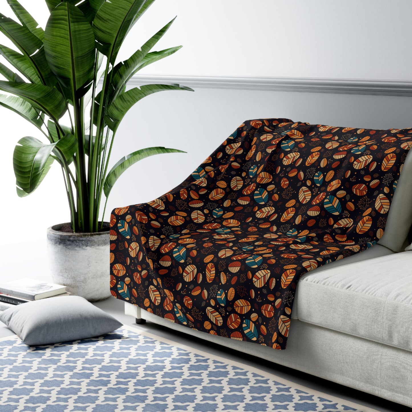 A throw blanket draped over a light gray sofa in a styled living room setting, featuring a dark brown background with a repeating pattern of orange, teal, and brown leaf motifs. A large green potted plant and a blue patterned rug are visible in the background.