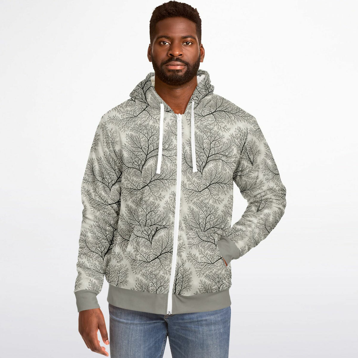 A man wearing a light gray zip-up hoodie with a black tree branch pattern, paired with blue jeans, photographed against a plain white studio background.