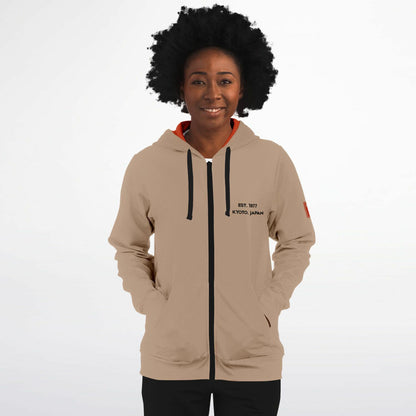 A woman with an afro hairstyle wearing a beige zip-up hoodie with black drawstrings, a black zipper, and a red inner hood lining. The hoodie features black text on the left chest reading 'est 1977 KYOTO JAPAN' and a small red patch on the right sleeve.