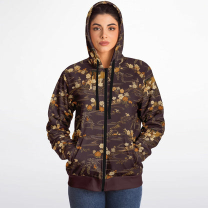 A woman wearing a dark brown hooded zip-up jacket with a beige and gold floral pattern, paired with blue jeans, photographed against a plain white studio background.
