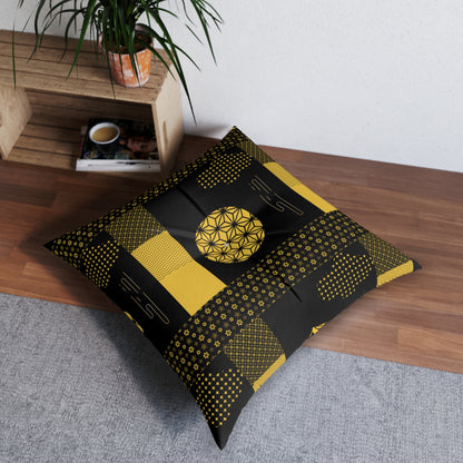 A square floor pillow with a black and yellow geometric patchwork pattern, styled on a gray rug on a wooden floor next to a wooden shelf with a plant.