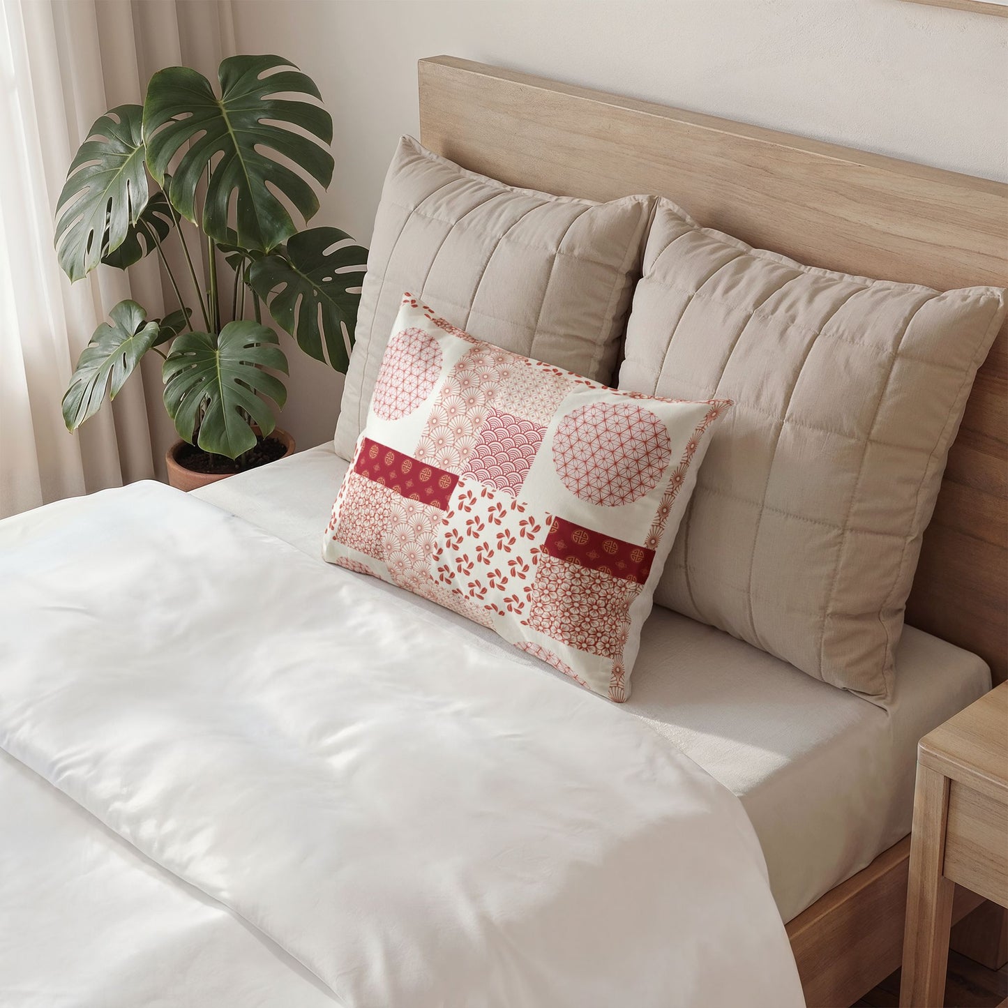 A decorative rectangular throw pillow with a red and cream patchwork pattern sits on a bed with white bedding, flanked by two large beige quilted pillows against a light wood headboard. A potted monstera plant is visible in the background.