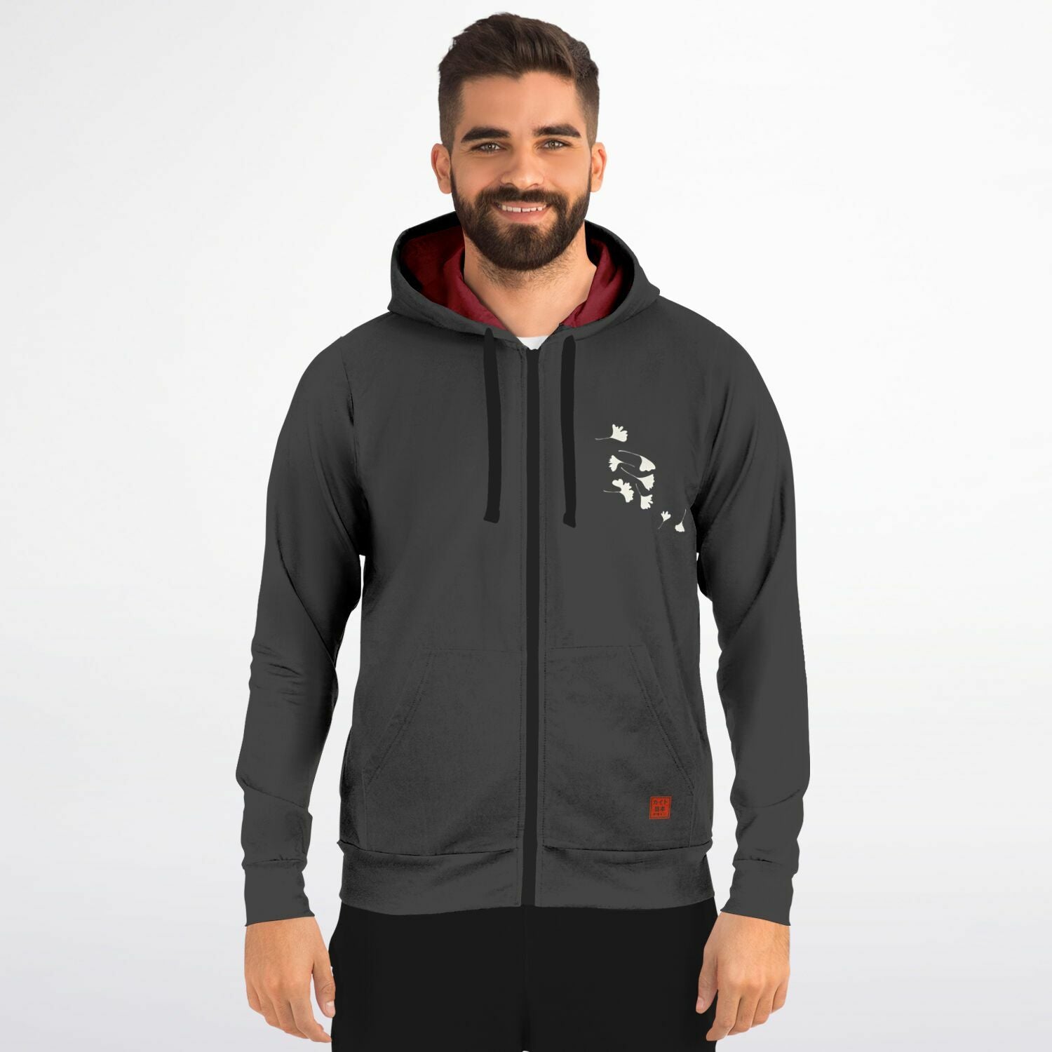 A man wearing a dark gray zip-up hoodie with a black drawstring, a white graphic print on the left chest, and a red-lined hood, standing against a light gray background.