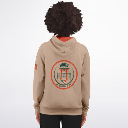 A person shown from the back wearing a beige hoodie with a large circular 'Kaito Volleyball Club' graphic on the back and a small orange patch on the sleeve, against a plain light gray studio background.