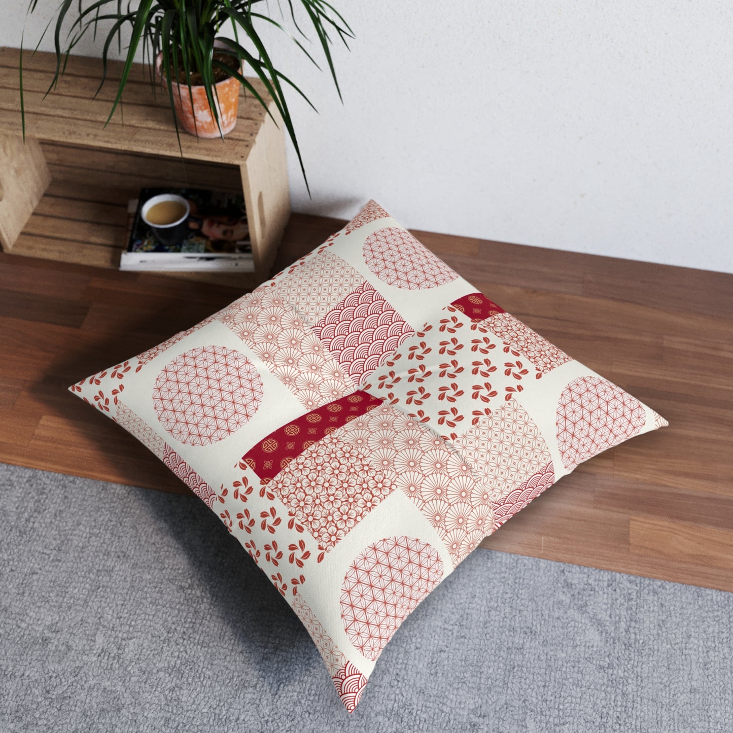 A square decorative throw pillow with a patchwork of Japanese-style patterns in pink, red, and cream, styled on a gray rug on a wooden floor next to a wooden shelf with a plant.