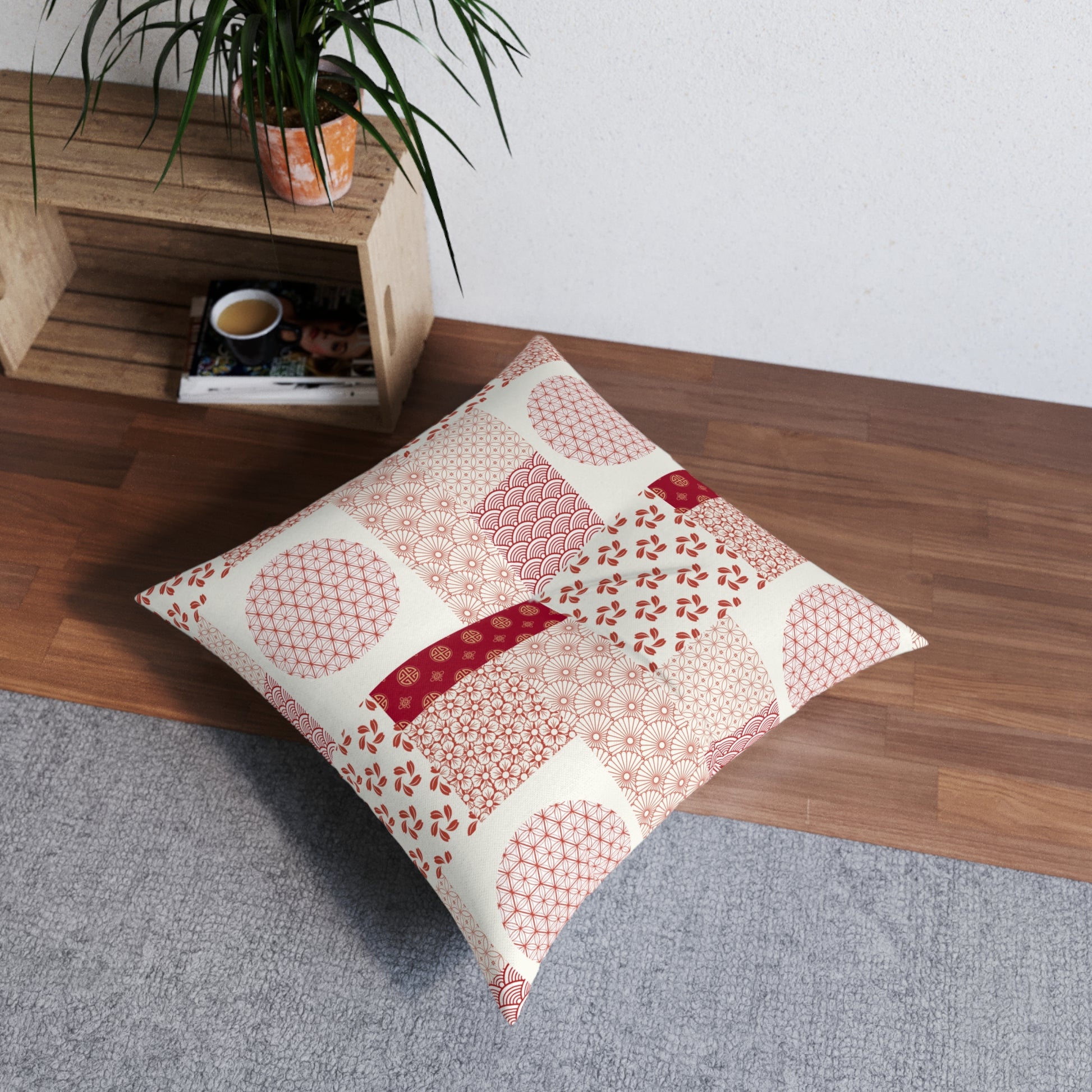 A square decorative throw pillow with a patchwork of red and pink Japanese-style patterns on an off-white background, styled on a gray rug in a room corner next to a wooden shelf with a plant.