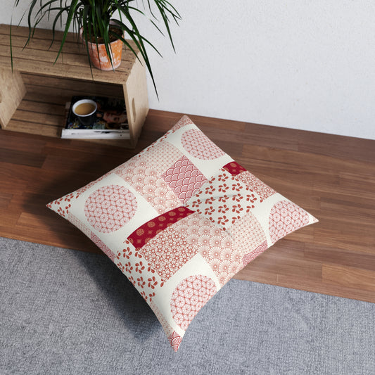 A square decorative throw pillow with a patchwork of red and pink Japanese-style patterns on an off-white background, styled on a gray rug in a room corner next to a wooden shelf with a plant.