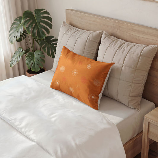 An orange decorative throw pillow with subtle white floral patterns rests on a bed with white bedding, flanked by two beige quilted pillows against a light wood headboard, with a green monstera plant in the background.